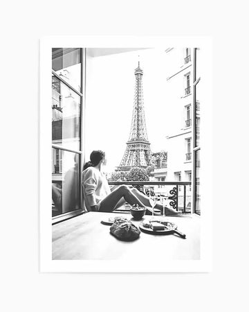 Framed black and white photograph of a woman sitting on a balcony overlooking the Eiffel Tower in Paris. The woman, seen from behind, wears a light-coloured top and looks out towards the iconic landmark. On the table in front of her are a loaf of bread, a bowl, and a plate with cheese and two wine glasses, suggesting a meal or picnic. The composition captures a serene moment with a classic Parisian view.