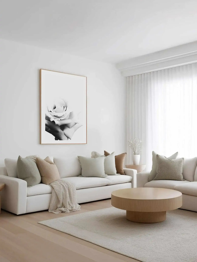 Black and white photographic art print featuring a close-up of hands holding a delicate rose bloom. The image has soft, artistic focus with subtle gradient tones and intimate composition. Framed in natural wood with white border, displayed on white wall above cream linen sofa styled with neutral throw pillows and blanket in a minimalist living room with light wood round coffee table and sheer white curtains.