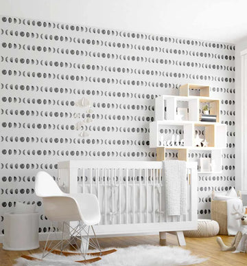 Nursery:
"Moon Phases wallpaper displayed on the back wall of a modern nursery. The wallpaper features a repeating pattern of hand-drawn moon phases in charcoal grey on a crisp white background, arranged in horizontal rows. The room includes a white crib with white bedding, a white rocking chair, a white mobile with cloud shapes, a white and natural wood shelving unit with books and plants, a white fluffy rug, and a woven basket with a stuffed reindeer toy."