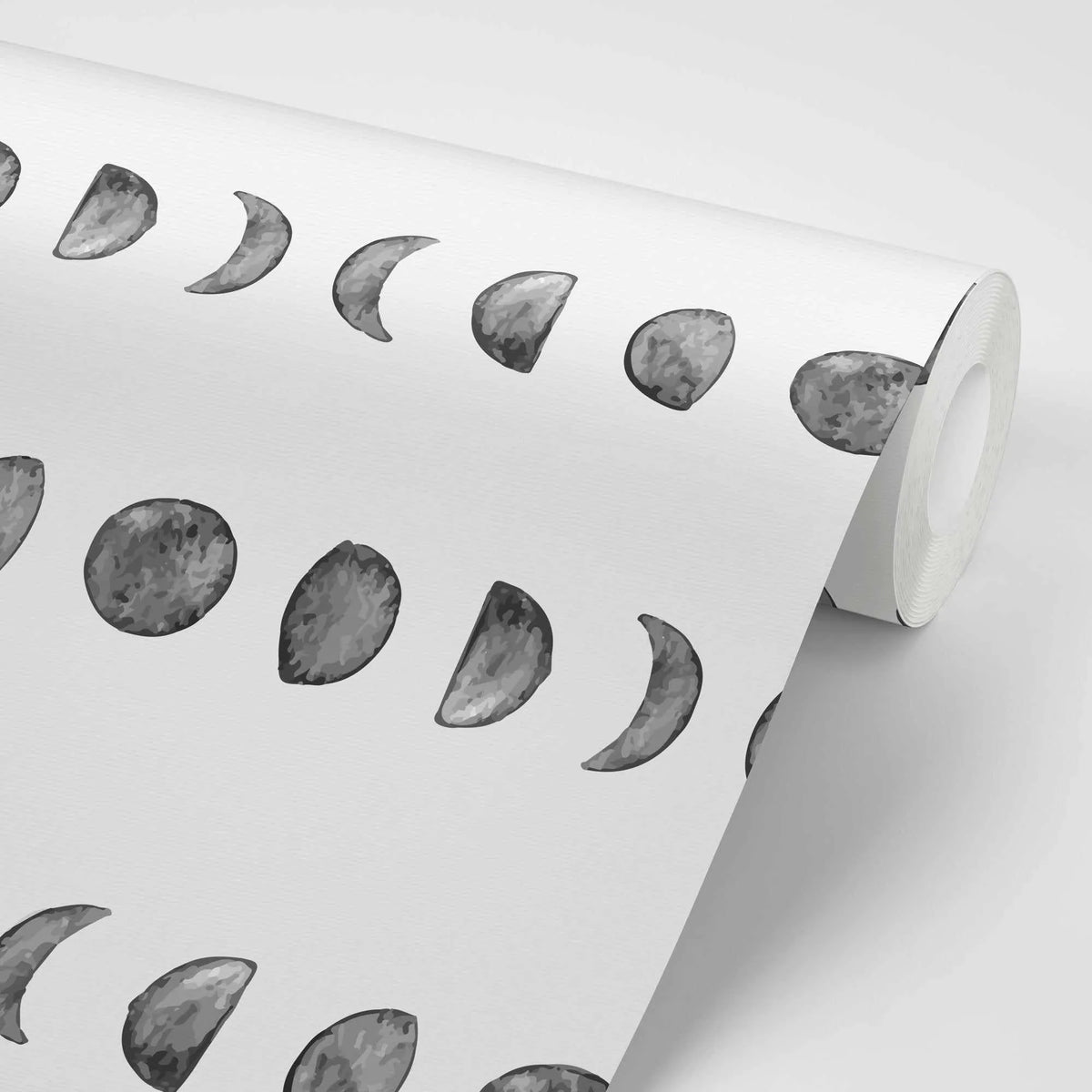 Peel and stick wallpaper featuring a repeating pattern of hand-painted moon phases in varying shades of grey on a white background, with a visible watercolor texture. The wallpaper is shown unrolled, with the right side curling into a roll.