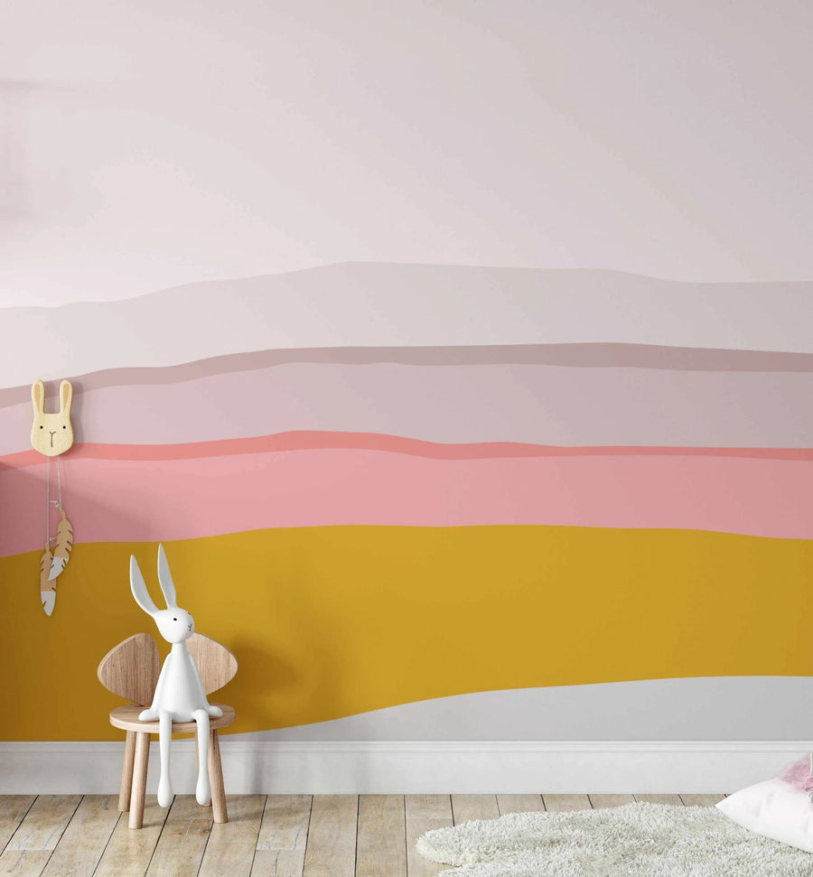 Children's Room:
Peel and stick wallpaper mural featuring an abstract landscape design with horizontal bands of colour in varying widths and wavy edges, creating a layered effect. The colours include pale pink, dusty rose, coral pink, and mustard yellow, with a thin white band at the bottom. A light wood bunny-shaped clock with feather pendulums hangs on the left, and a white bunny figurine sits on a small wooden chair in the foreground. The room has light wood flooring and a fluffy white rug.