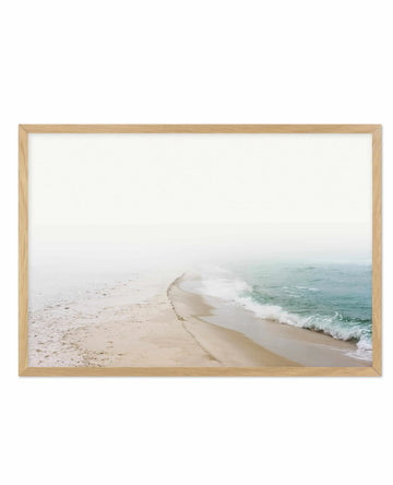 Framed photography art print of a misty beach scene with a natural oak frame and white border. The composition features a sandy beach with gentle waves breaking on the shore, with the ocean and sky fading into a soft white mist.
