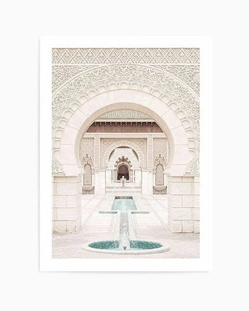 Framed photography art print featuring a symmetrical architectural scene of a white Moroccan courtyard with intricate carvings and arches, leading to a series of smaller arches and a central fountain with turquoise water. The print has a white border.