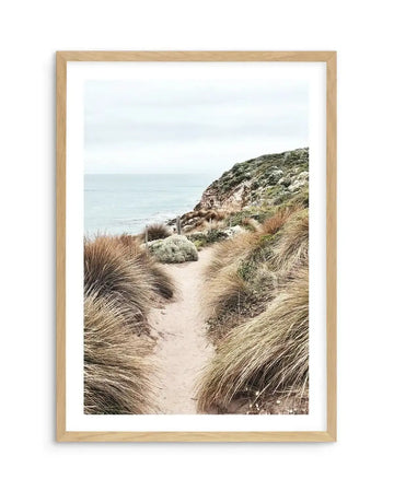 Framed photography art print of a coastal path with a natural wood frame and white border. The composition features a sandy path winding through tall, dry grasses and low-lying green and grey-green shrubs, leading towards a rocky cliff face and the ocean under an overcast sky.