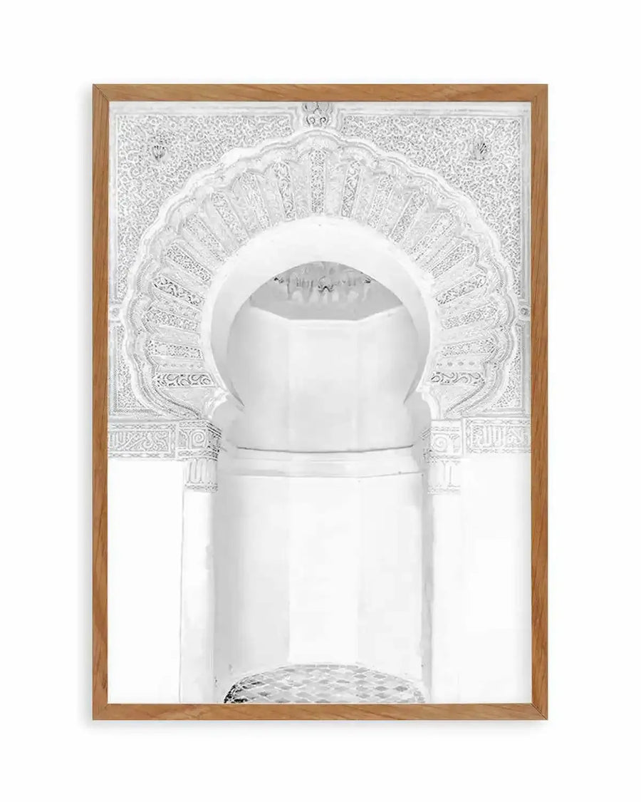 Framed black and white photograph of a Moroccan arch with intricate carved details, displayed in a natural oak frame with white border. The arch features a horseshoe shape with elaborate floral and geometric patterns, leading into a recessed alcove with a textured floor.