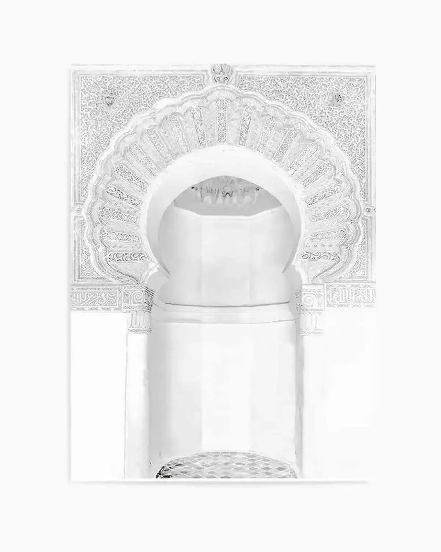 Monochromatic art print featuring a detailed Moroccan arch in shades of white and light grey, with intricate geometric and floral patterns carved into the surrounding wall and the arch itself. The arch has a scalloped, fan-like design at its top, leading into a recessed, multi-faceted interior. The lower part of the image shows a textured, possibly tiled, surface at the base of the arch. The overall impression is one of delicate craftsmanship and architectural elegance.