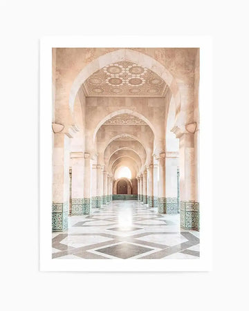 Framed photograph of a long, arched hallway in a Moroccan mosque, with a warm, bright light at the far end. The hallway features light beige stone arches and columns, with intricate geometric patterns carved into the ceilings and the upper sections of the arches. The lower half of the columns and walls are adorned with green and white patterned tiles. The floor is a polished white marble with a repeating dark grey geometric pattern, reflecting the light from the end of the hall.