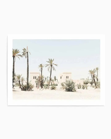 "Photography art print featuring a desert landscape with light sand and sparse vegetation under a pale blue sky. The composition includes several tall palm trees and two light-colored buildings with arched windows, evoking a serene, minimalist Moroccan scene."
