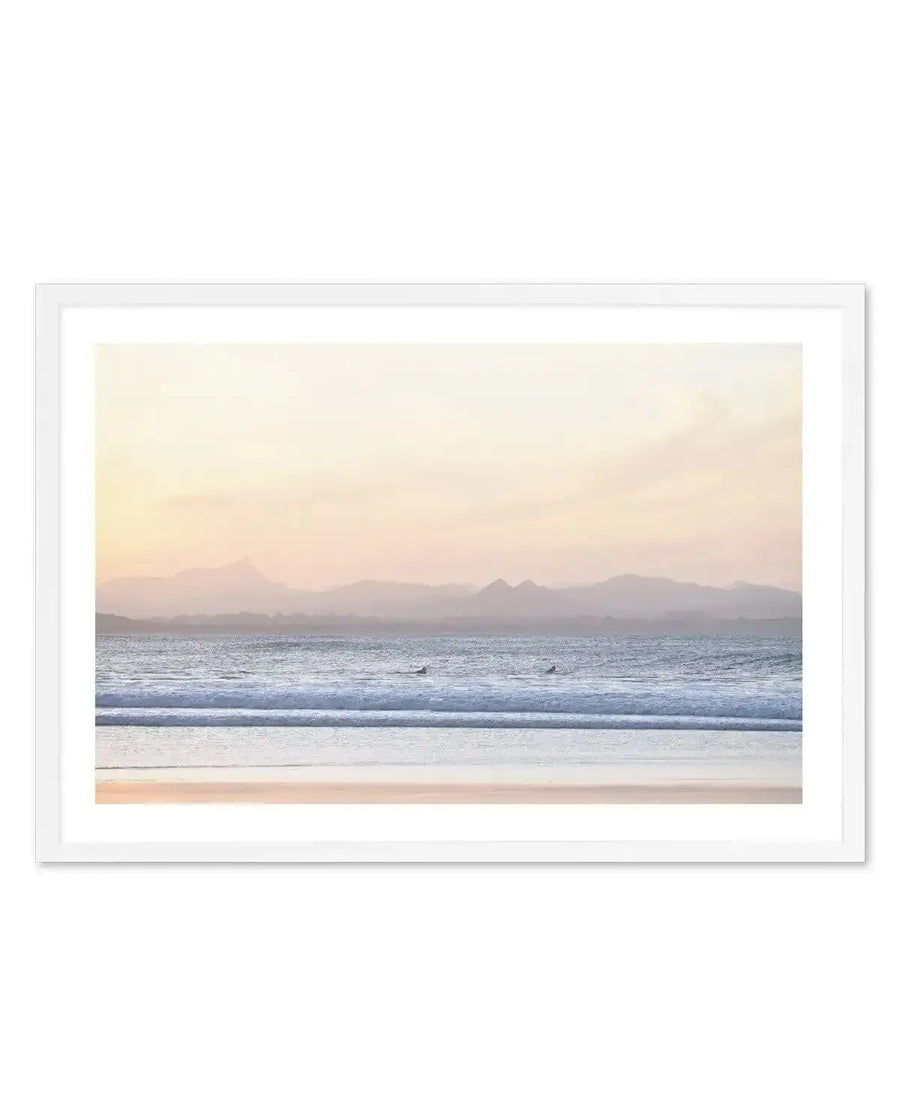 Framed art print with white border showing a serene coastal landscape at dawn or dusk. The composition features a sandy beach in warm peachy tones in the foreground, gentle waves with white foam in soft blue-grey tones in the middle ground, and misty mountains silhouetted against a pale yellow and blush pink sky across the horizon. Two small figures appear to be surfing in the water. The overall mood is calm and atmospheric with soft, muted colours. The print is displayed in a white wooden frame.