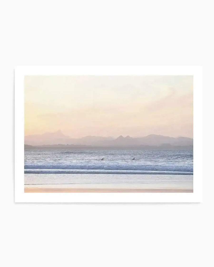 Serene coastal landscape art print featuring a misty dawn view of Mount Warning rising in soft purple and blue hues across the ocean horizon. Sandy beach in warm beige tones stretches across the foreground, with gentle waves in pale blue and white rolling onto the shore. Soft peachy and cream tones blend across the sky. The photograph-style print is mounted with a clean white border on a white background, capturing a calm, minimalist aesthetic of a surfer's viewpoint at Byron Bay.