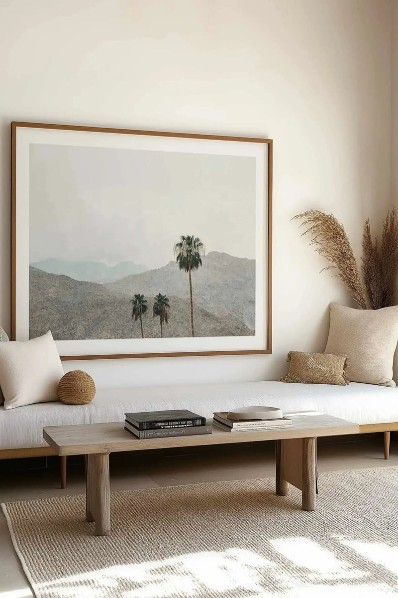 Framed art print displayed on a white bedroom wall above a light wood bench. The photograph shows three palm trees against layered desert mountains in muted greens and grays under a hazy sky, creating a serene landscape. The print has a warm wood frame with a white border. Below, a natural wood coffee table holds stacked books, and cream-colored pillows with woven textures and pampas grass complete the minimalist, earthy interior setting.
