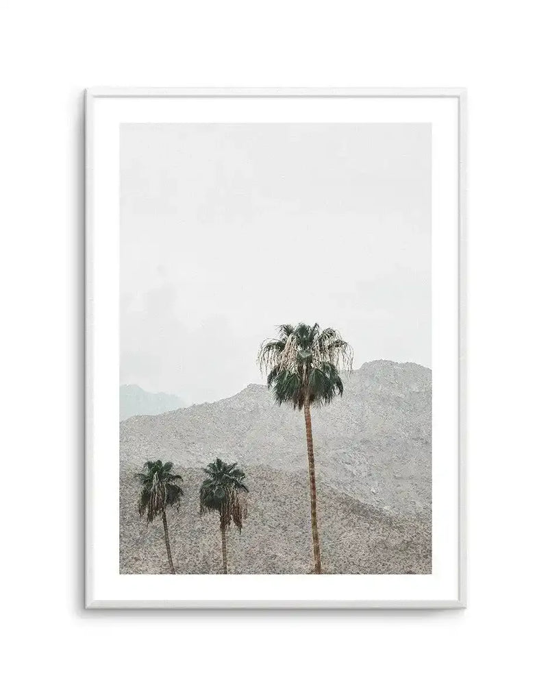 Framed art print featuring a minimalist desert landscape photograph. Three palm trees with thin trunks and wispy fronds stand against layered, rocky mountains in muted tan and grey tones. The sky is pale and overcast. The photograph has a soft, hazy quality with a calm, contemplative mood. The print is displayed in a white frame with white border matting.