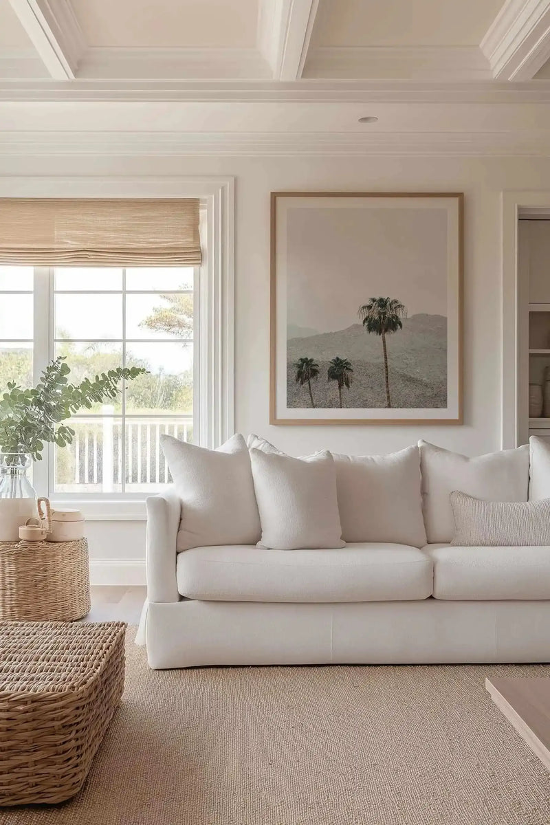 Framed art print depicting three palm trees against misty, sage-green mountains in muted tones. The photograph-style print is mounted in a natural wood frame with white border, displayed above a cream linen sofa in a bright, minimalist living room with coffered ceiling, large windows, woven accessories, and neutral furnishings.