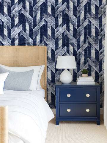 Lifestyle shot of a bedroom featuring navy herringbone wallpaper behind a rattan headboard. The wallpaper has a repeating pattern of dark indigo blue vertical stripes interspersed with white stripes, overlaid with a zigzag pattern of thin, textured white lines. The room includes a bed with white bedding and a light blue textured pillow, and a navy blue nightstand with a white lamp and stacked books.