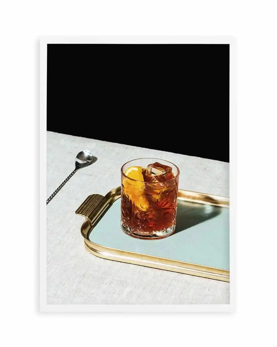 Framed photograph of a Negroni cocktail on a mirrored tray with a gold rim and a silver stirring spoon on a white linen tablecloth, with a dark, shadowy background. The cocktail is a deep amber color with ice cubes and an orange slice garnish.