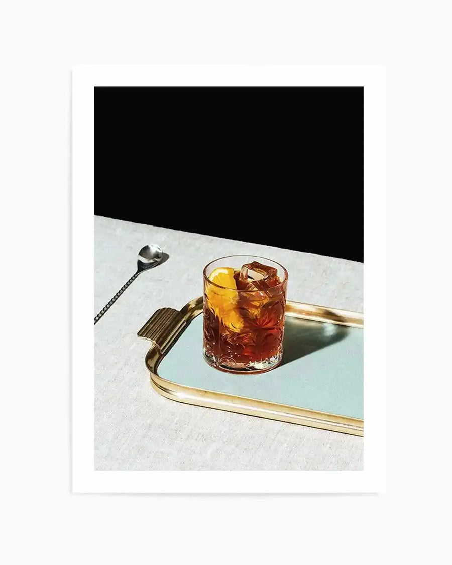 Framed photograph of a Negroni cocktail on a light blue tray with a gold rim, set on a textured light grey surface against a stark black background. The cocktail, a deep amber color with ice cubes and an orange slice, is presented in a clear, ornate glass. A long, twisted silver spoon rests on the grey surface to the left.
