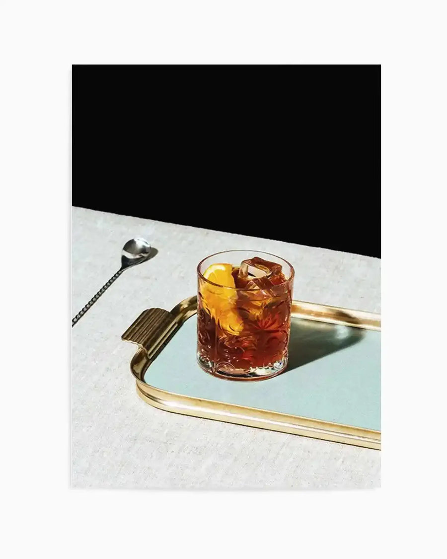 Framed photography art print featuring a Negroni cocktail in a textured glass with ice and an orange slice, resting on a light blue mirrored tray with a gold rim. A long, silver stirring spoon is visible to the left of the tray on a light grey textured surface. The background is a stark black, creating a dramatic contrast with the illuminated foreground.