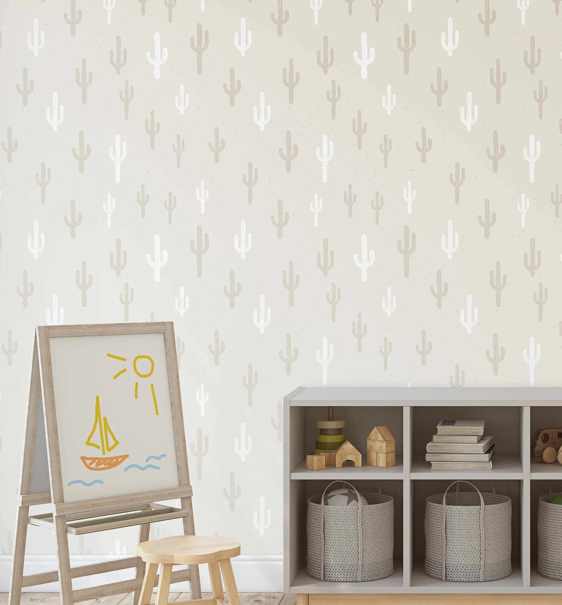 Lifestyle shot of a child's room featuring peel and stick wallpaper with a repeating pattern of hand-drawn saguaro cacti in alternating soft white and light taupe on a warm cream background with subtle speckle texture. The wallpaper covers a wall behind a light wood easel with a child's drawing of a yellow sailboat and sun, and a grey open shelving unit filled with toys and books.