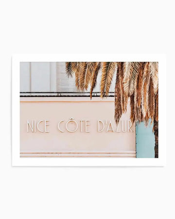"Photography art print featuring the words 'NICE CÔTE D'AZUR' in white raised lettering on a pale pink wall, partially obscured by the fronds of a palm tree. The composition captures a sunny, relaxed mood with a hint of a light blue wall or shutter visible on the right."