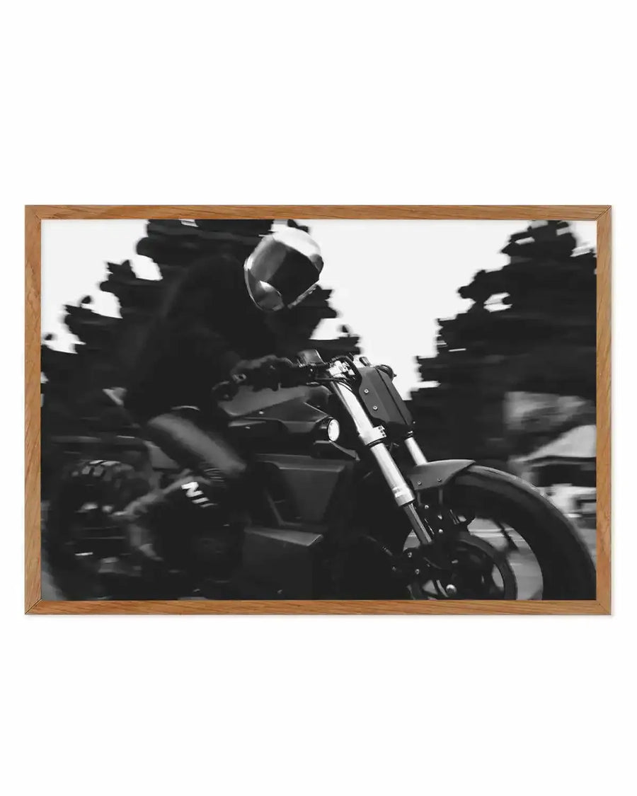 Framed black and white photograph of a person riding a motorcycle at speed, displayed in a natural oak frame with white border. The composition captures the rider in a helmet and dark clothing, leaning into a turn, with blurred trees in the background suggesting rapid motion.