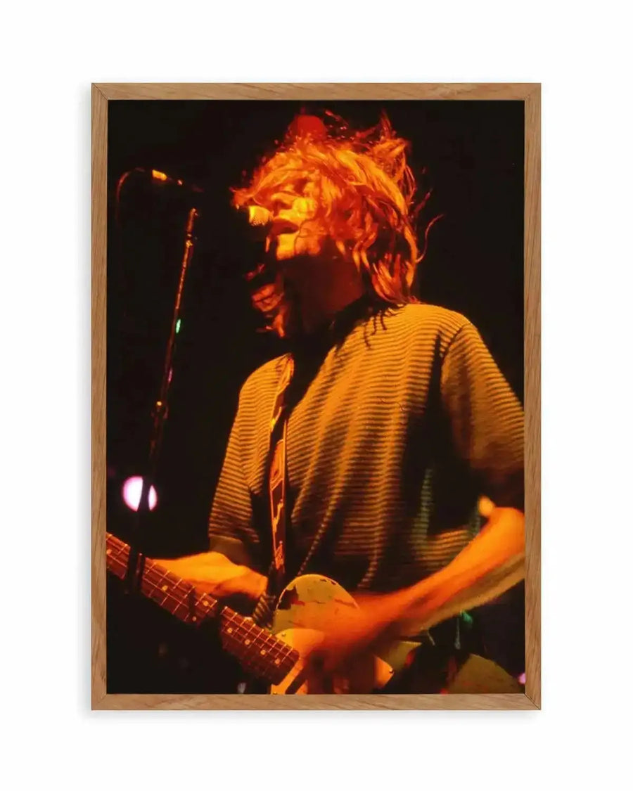 Framed photography art print of a musician on stage, captured in a dynamic moment with warm orange stage lighting. The musician, with long, light brown hair flying, is playing an electric guitar and singing into a microphone. They are wearing a striped short-sleeved shirt and a guitar strap. The background is dark, with subtle purple light visible on the left. The print is displayed in a natural oak frame with a white border.