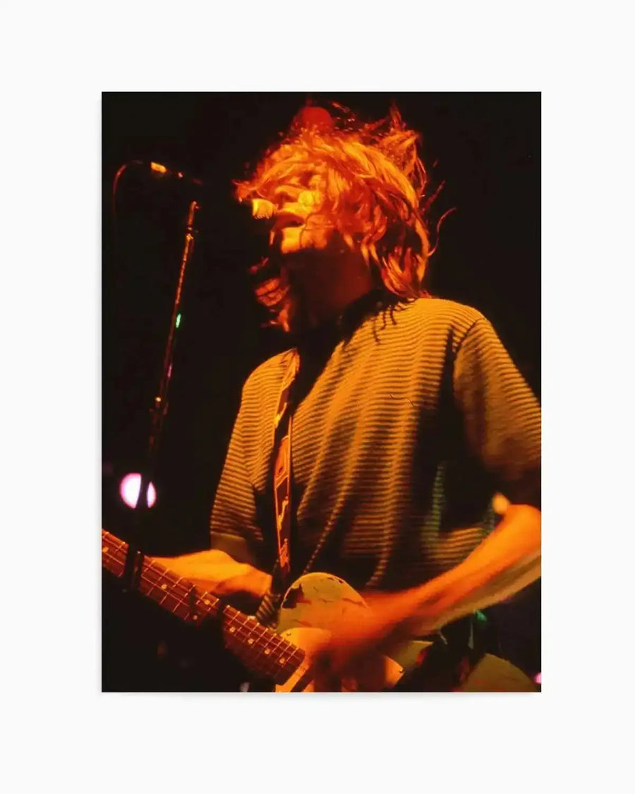 "Photography art print of Kurt Cobain performing live on stage, captured in warm orange and yellow light against a dark background. Cobain is shown from the waist up, playing a guitar, with his hair flying wildly as he looks upwards and to the right. He wears a striped short-sleeved shirt and a guitar strap. The image has a grainy, raw texture, conveying the energy of a live concert."