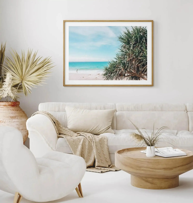 Framed art print displayed on a white wall above a cream linen sofa. The print shows a tropical beach scene with turquoise water, pale sand, and dark green palm fronds framing the right side. A lone figure wades in the shallow water under a partly cloudy blue sky. The print has a natural wood frame with a white border. The room features coastal-inspired décor including a curved white armchair, wooden round side table, woven basket with dried grasses, and a beige throw blanket.