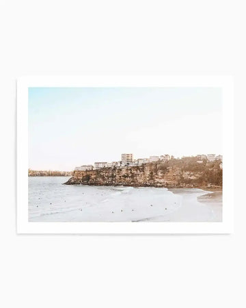 Framed photograph of a coastal beach scene with a white border. The composition features a wide, light sandy beach with gentle waves and several surfers in the water, backed by a rocky cliff face topped with white buildings under a pale blue sky.