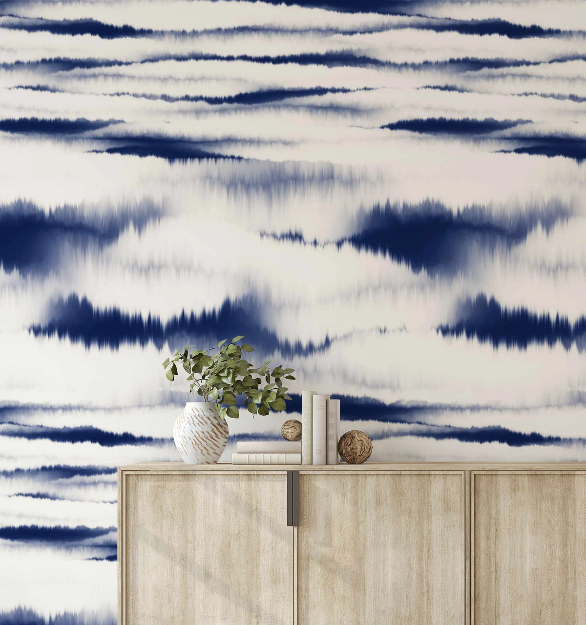 "Nobu Shibori wallpaper featuring a repeating pattern of horizontal indigo blue and white stripes with a blurred, tie-dye effect, creating a dynamic, organic design. The wallpaper is displayed in a living room above a light wood console table, which is adorned with a white ceramic vase holding green foliage, a stack of books, and two decorative wooden spheres."