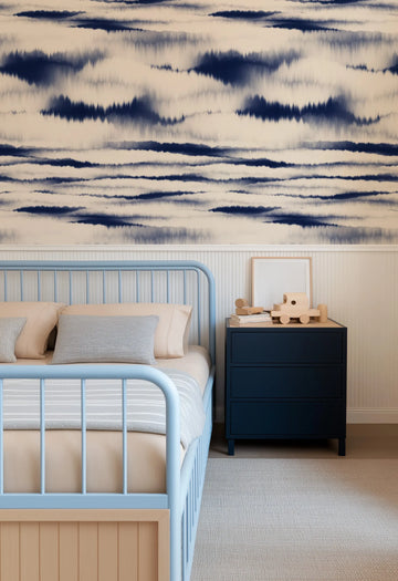 "Nobu Shibori wallpaper with horizontal bands of deep indigo blue and pale grey, resembling abstract tie-dye or ink wash, covering the upper wall of a child's bedroom. The wallpaper is displayed above white vertical beadboard paneling. A light blue metal bed frame with beige bedding and a grey striped blanket is in the foreground, with a dark blue nightstand and wooden toys to the right."