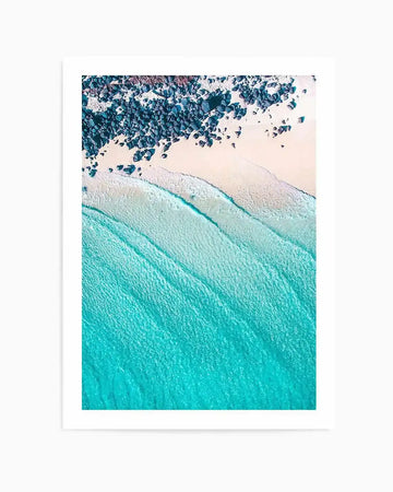 Aerial photography art print of Noosa beach, featuring a vibrant turquoise ocean with gentle waves meeting a pale sandy beach scattered with dark blue-grey rocks, printed on fine art paper with a matte finish.