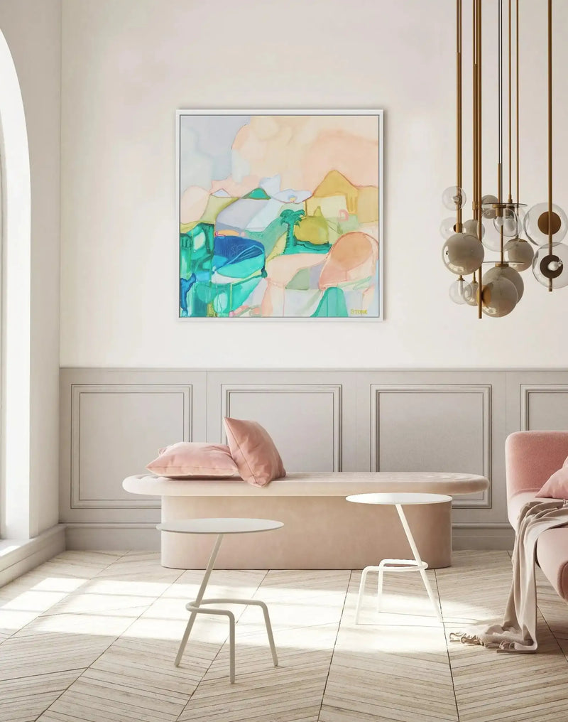 Lifestyle shot of a minimalist living room featuring a large framed abstract art print above a curved blush pink velvet bench. The print, titled "Noosa Palm," features an abstract landscape with organic shapes in shades of teal, blue, sage green, peach, and cream, with a white frame and white border. The room has light grey wainscoting, a light wood chevron floor, and a modern gold and glass chandelier. Two small white side tables and a pink velvet sofa with a cream throw are also visible.