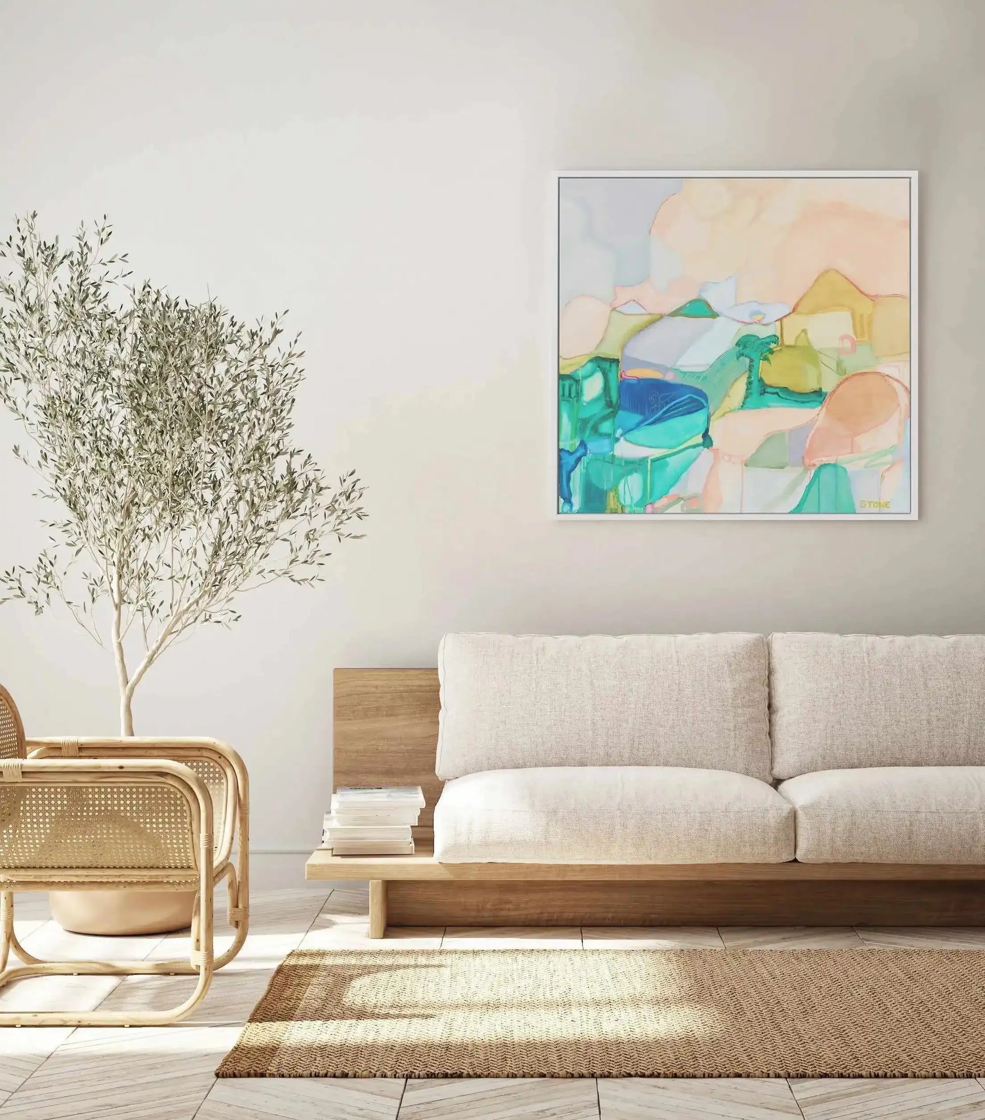 Lifestyle shot of a minimalist living room featuring a framed abstract canvas print above a light cream sofa. The print, titled "Noosa Palm," features an abstract landscape with organic shapes in shades of teal, aqua, soft peach, and muted yellow, suggestive of rolling hills and water, with a white frame. The room includes a light wood sofa base, a stack of books, a large potted olive tree, and a rattan chair on a light-coloured tiled floor with a natural woven rug.