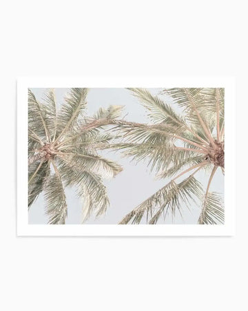 "Art print featuring a low-angle photograph of two palm trees against a pale blue sky. The fronds are a muted sage green with hints of dusty rose on the undersides, creating a soft, serene tropical composition."