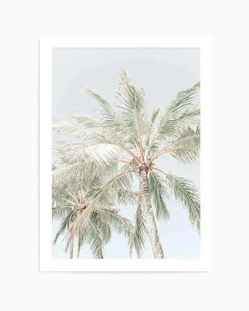 Art print featuring a pale, high-key photograph of two palm trees against a soft, light blue sky. The palm fronds are a muted sage green and cream, with visible texture in the leaves and trunks, creating a serene, minimalist tropical aesthetic.