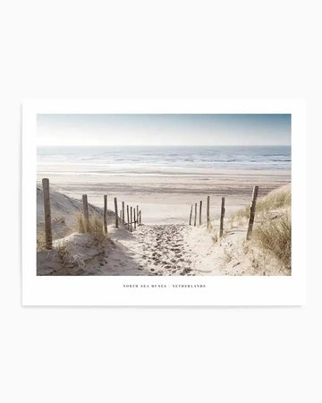 Framed photograph of a coastal beach scene with a path leading through sand dunes to the ocean, with a white border. The composition features a sandy path with footprints, flanked by wooden posts and dry grass, leading towards a wide expanse of beach and the calm, light blue sea under a bright sky.
