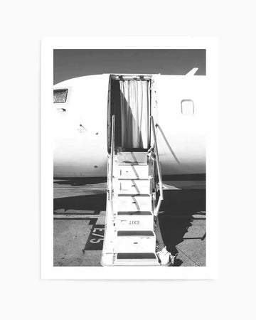 Framed black and white photograph of an airplane boarding ramp with the door open. The composition shows the white side of the aircraft with a window and a small fin, and the white boarding stairs with handrails leading up to the open door, which has a curtain visible inside. The word "EXIT" is visible on one of the steps, and "E/5" is painted on the tarmac next to the stairs.