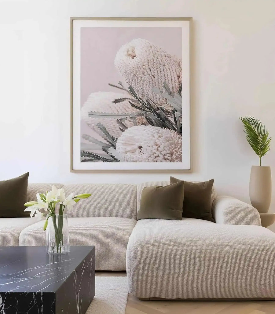 Large framed art print displayed above a textured cream sectional sofa in a minimalist living room. The print features a close-up photograph of three white Banksia flowers with intricate, textured blooms and spiky green leaves against a soft blush pink background, displayed in a natural wood frame with white border. The room includes dark olive green throw pillows, a black marble coffee table with white veining, and a tall cream vase with a single green palm frond.