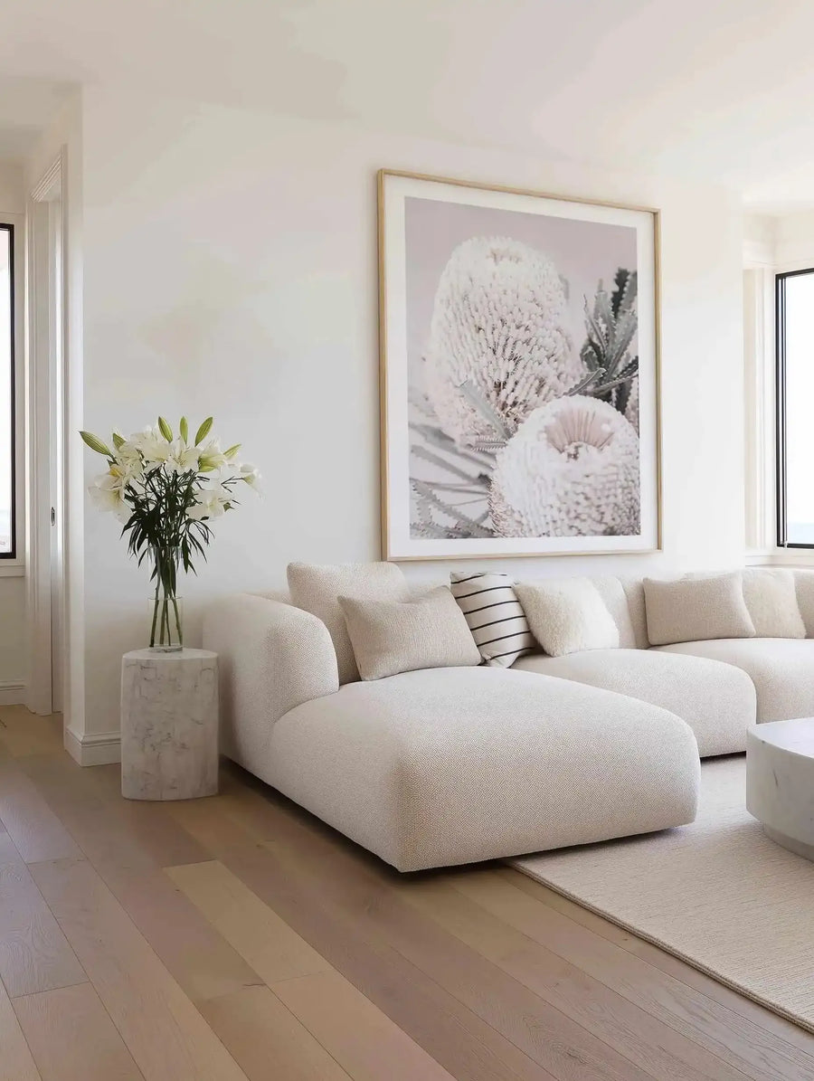 Large framed art print displayed above a curved cream sofa in a minimalist living room. The print features a close-up photograph of three white Banksia flowers with textured, spiky petals and green foliage against a soft grey background, with a natural wood frame and white border. The room includes a white marble side table with a vase of white lilies and a cream rug on a light wood floor.