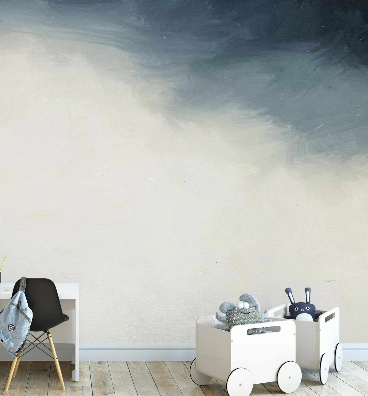 Lifestyle shot of a child's room featuring Nuit peel and stick wallpaper. The wallpaper has a hand-painted, abstract design with broad, horizontal brushstrokes that transition from a deep, dark blue at the top to a soft, creamy white at the bottom, creating a gradient effect. The room includes a white desk with a black chair and a light blue backpack, and two white toy storage carts filled with plush toys on a light wood floor.