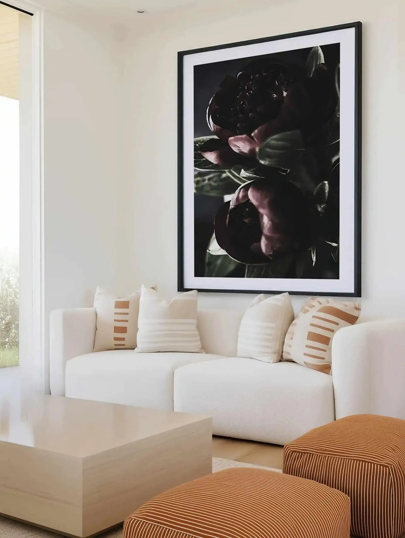 Black-framed art print displaying dark moody photograph of deep burgundy and black peonies with green stems against dark background, mounted with white border above cream upholstered curved sofa styled with cream and tan striped throw pillows in bright minimalist living room with light wood coffee table and striped ottoman in foreground.