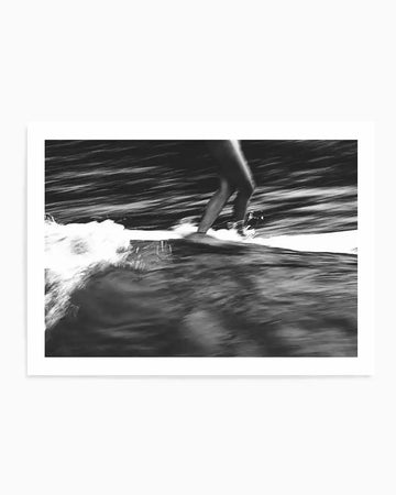 Framed black and white photograph capturing a surfer's legs in motion on a surfboard, riding a wave. The image has a blurred, dynamic quality, suggesting speed and movement, with the dark water streaking in the background and white foam breaking around the board.
