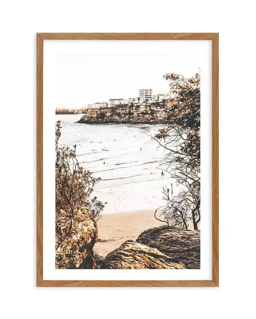 Framed photography art print of a coastal beach scene with a natural wood frame and white border. The composition captures a wide sandy beach with gentle waves and a few people in the water and on the sand, viewed from a slightly elevated perspective between two rocky outcrops with sparse, dry-looking vegetation. In the background, a cliffside is topped with buildings under a bright, clear sky.
