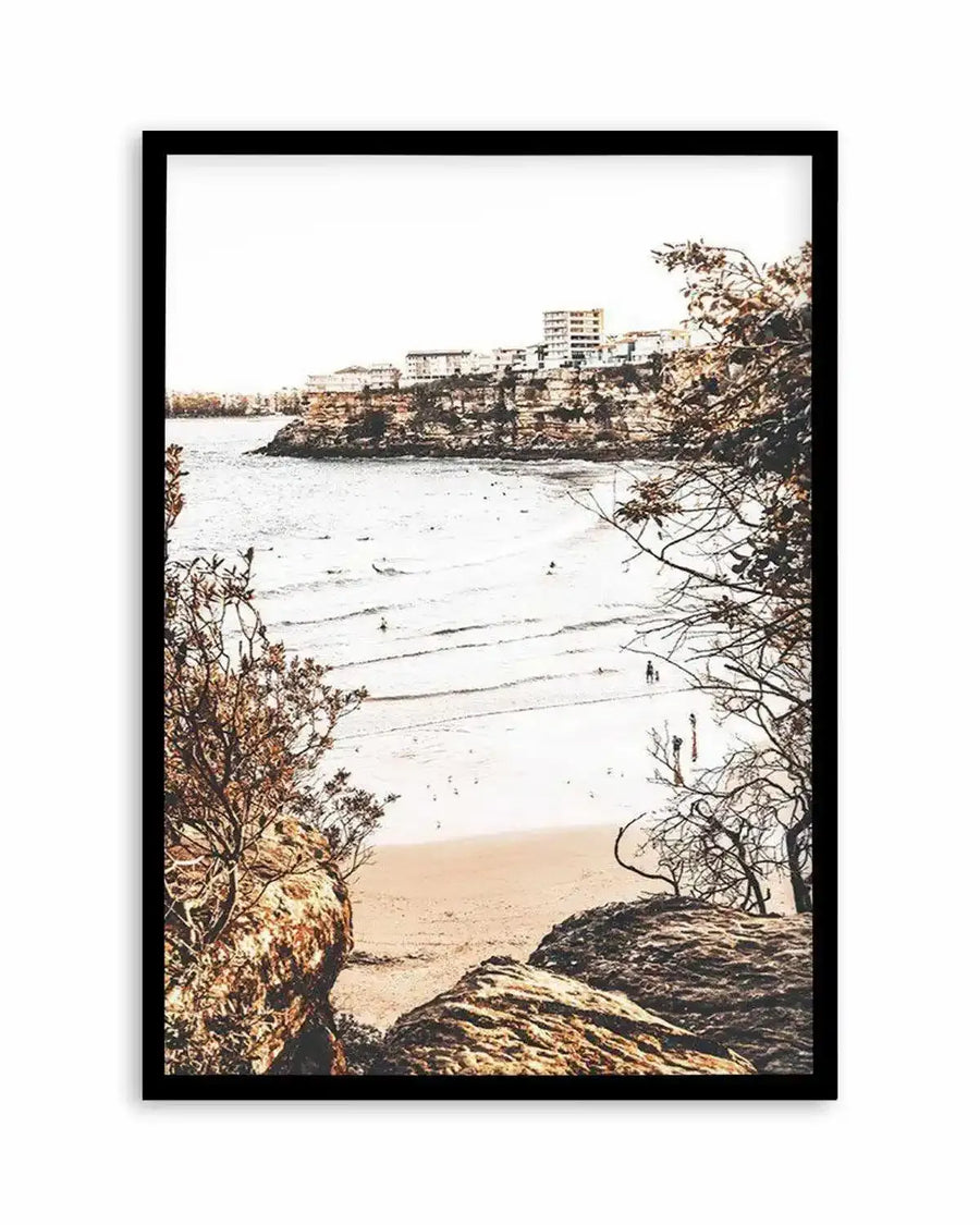 Framed art print featuring a high-contrast photograph of a coastal beach scene with a black frame and white border. The composition captures a wide sandy beach with gentle waves, a few people walking and surfing, and a rocky headland with buildings in the background. The foreground features dark, textured rocks and bare trees with brown leaves, framing the view of the beach.