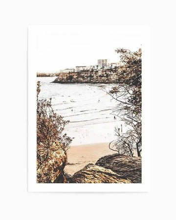 Framed photograph of a coastal beach scene with a white border. The composition features a wide sandy beach with gentle waves and several people swimming and walking, framed by dark brown trees and rocks in the foreground, and a distant cliffside town under a bright, clear sky.