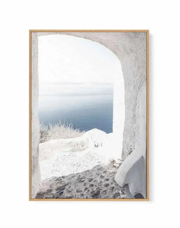 Framed canvas art print featuring a photograph taken through a white-walled arched doorway looking out onto a snow-covered landscape and calm blue ocean. The composition shows textured white and grey stone walls framing the rounded archway, with frost-covered vegetation visible in the middle ground and a serene seascape beyond. The image has a cool color palette of whites, greys, and blues with soft natural lighting. The canvas is mounted in a natural wood frame with a white border.