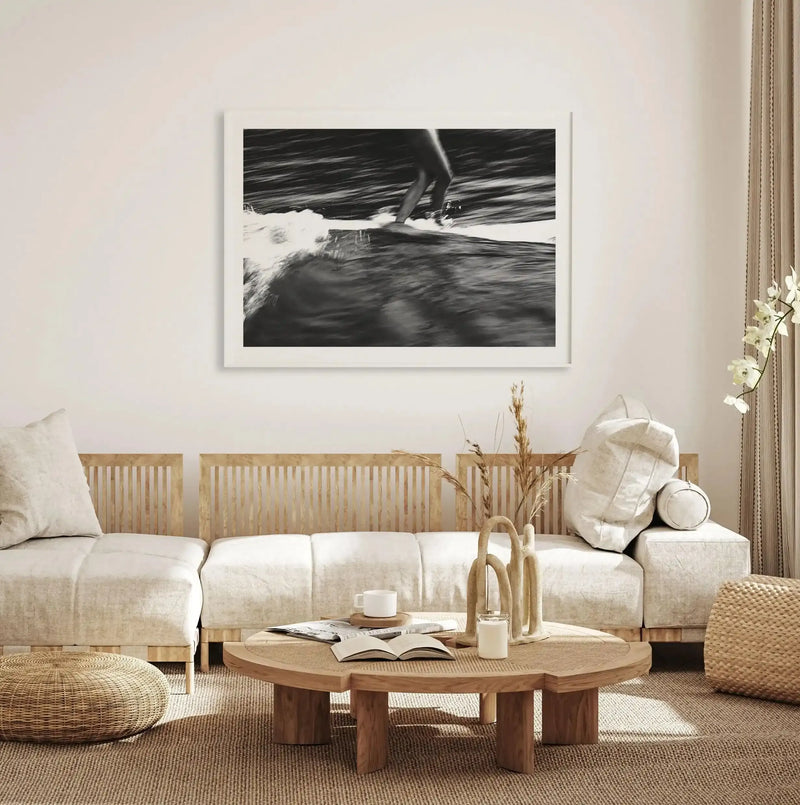 Lifestyle shot of a minimalist living room featuring a large framed black and white photograph of a surfer riding a wave. The art print is displayed above a cream sectional sofa with light wood slatted backs. A round wooden coffee table with a woven cane top sits on a textured natural fiber rug, adorned with books, candles, and dried pampas grass in a vase. A woven ottoman is visible in the foreground.
