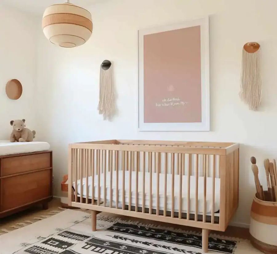 Framed art print with dusty rose background and white frame, displaying the text "oh darling, but what if you fly" in light cream-colored lettering, hung on a cream wall above a natural wood crib in a minimalist nursery room with woven wall hangings, a pendant light, wooden dresser, and patterned area rug.