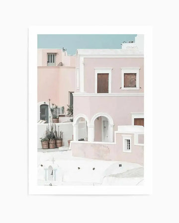 Framed art print depicting a minimalist architectural scene of whitewashed and dusty rose-colored buildings with arched doorways and small windows, set against a turquoise sky. The illustration uses soft watercolor tones and includes potted plants and muted earth-toned accents. Presented in a white frame with white border, evoking Mediterranean island architecture.