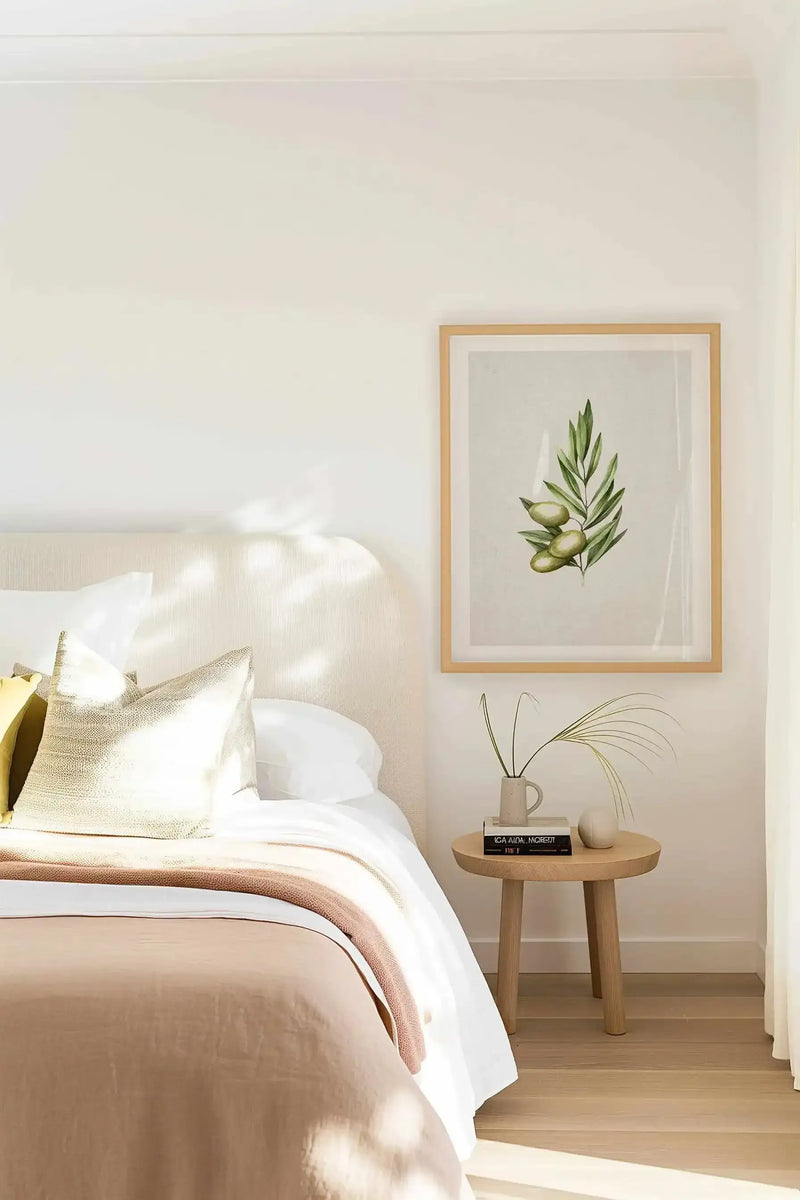 Framed art print featuring an olive branch with three green olives and slender green leaves, displayed above a light beige upholstered headboard in a bright, minimalist bedroom. The print has a natural wood frame and white border. A light wood bedside table holds books and two small vases with dried grasses. The bed is dressed with white sheets, a light brown blanket, and textured cream pillows.